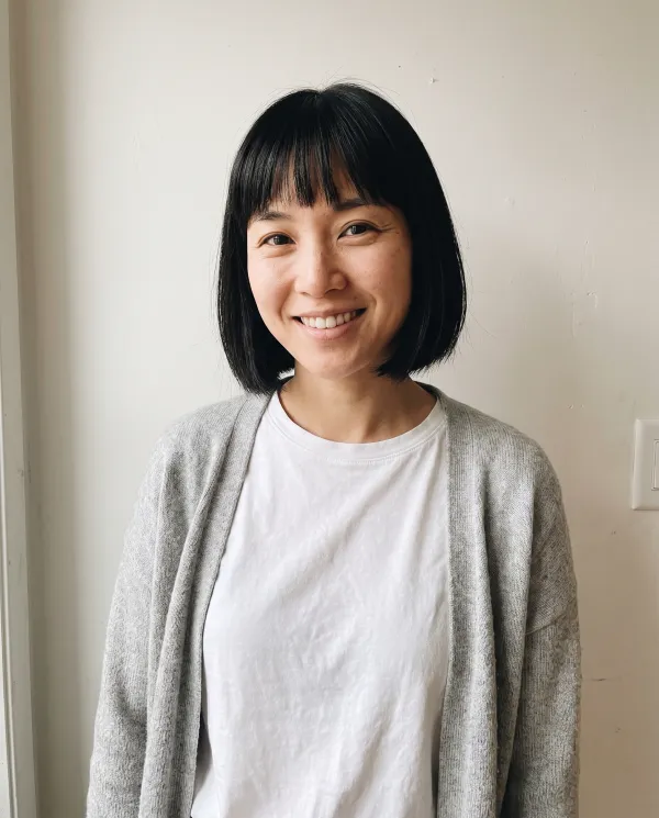A woman with a black bob and bangs in a grey cardigan against a white wall