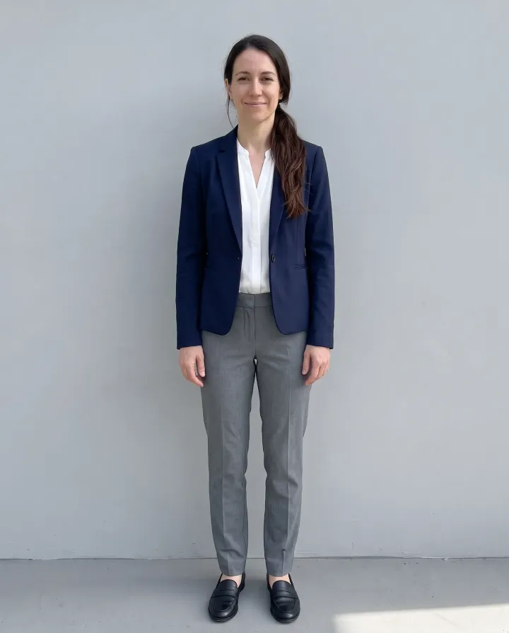 a woman in her early 30s with a long dark ponytail, standing against a light grey wall, in a navy blazer, white blouse and grey trousers