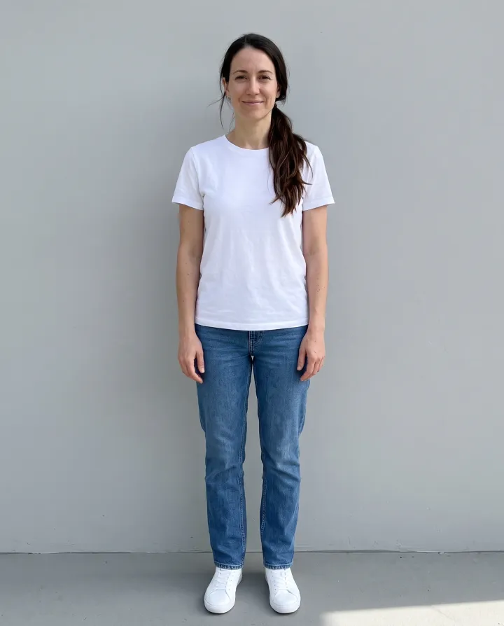a woman in her early 30s with a long dark ponytail, standing against a light grey wall, in a white t-shirt, jeans and sneakers