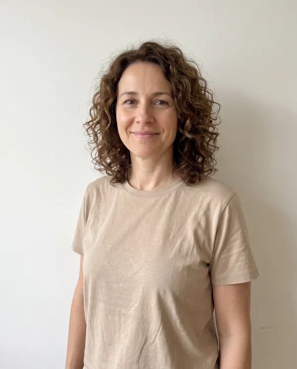 A woman with curly brown hair in a plain beige t-shirt against a light wall