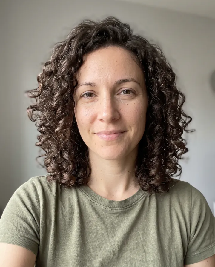 A woman with shoulder-length curly dark brown hair in an olive t-shirt
