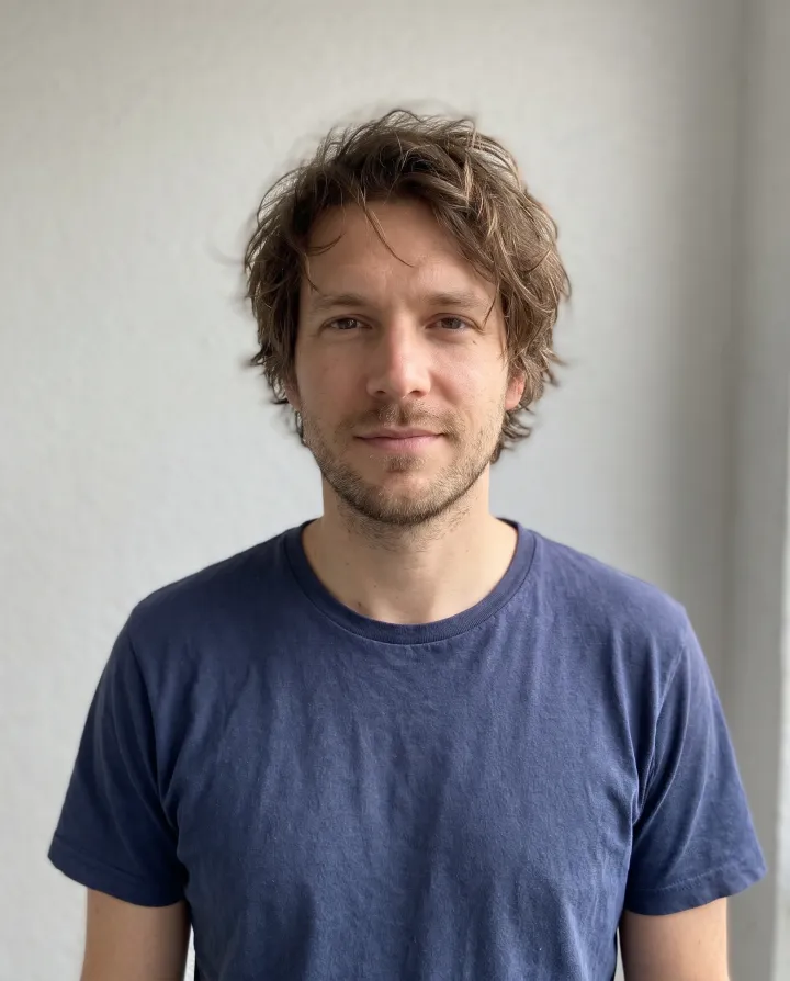 A man with medium-length messy brown hair in a navy t-shirt against a light wall