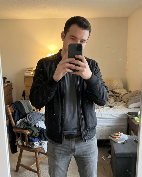 A mirror selfie of a man in a black bomber jacket in a messy bedroom