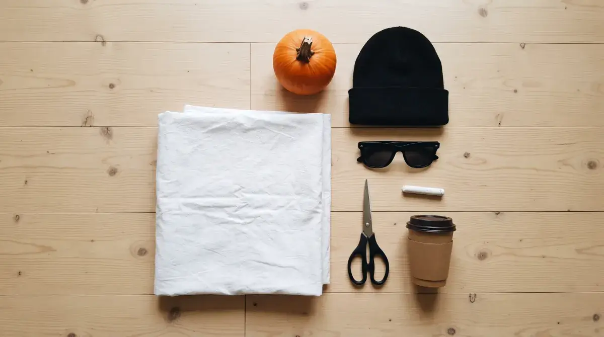 A white bed sheet, chalk, scissors, a black beanie, sunglasses, a coffee cup and a small pumpkin laid out on a wooden floor