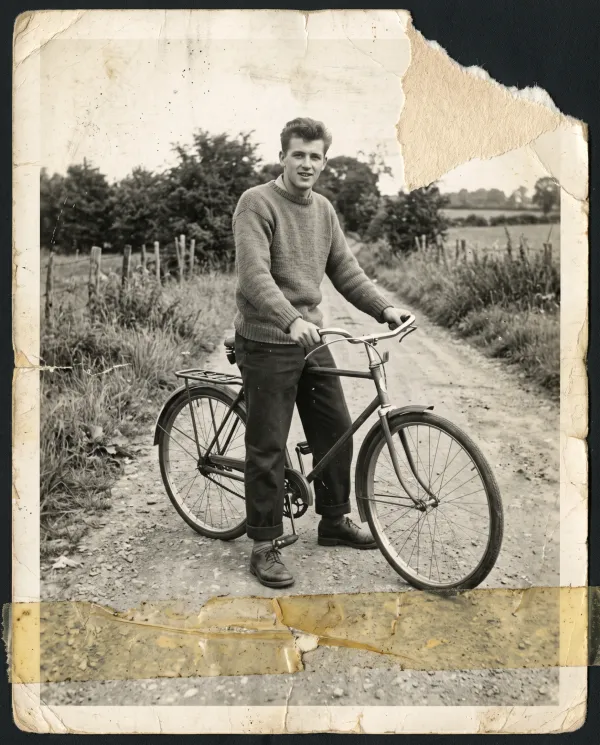 A black-and-white photo of a young man with a bicycle, the top corner torn off and tape across it