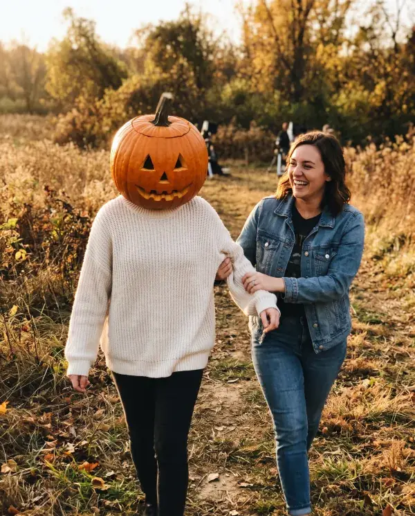 A friend guiding a woman wearing a real carved pumpkin head along a path in an autumn field